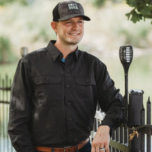 Man wearing black Crossroad Long Sleeve shirt outside leaning against a fence. 