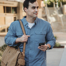 Man wearing Saddle blue Crossroad Long Sleeve shirt walking outside with saddle color bag over his shoulder and holding his phone. 