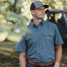 Man wearing slate blue Crossroad Short Sleeve Shirt while standing outside in the woods. 