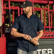 Man wearing deep blue Craftsman Short Sleeve Shirt standing outside in front of a cherry picker. 