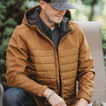 Man wearing saddle heather Pinnacle Puffer Jacket while staring a fire outside