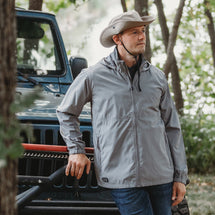 Man wearing grey River jacket standing outside leaning against car.